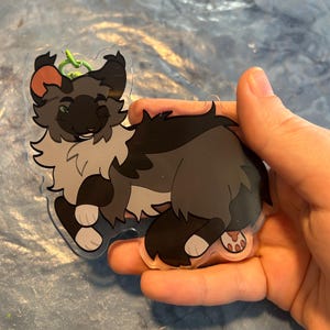 Pu&ograve; includere: Un portachiavi in acrilico trasparente con un design a forma di gatto dei cartoni animati. Il gatto ha pelo grigio, nero e bianco con un tocco di arancione sull'orecchio. Il portachiavi ha un anello verde.