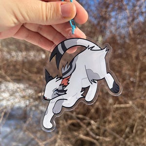 Op de afbeelding: Een heldere acryl bedel met een gestileerde witte en grijze kat in een rennende pose. De kat heeft zwarte en rode accenten en is bevestigd aan een blauwe metalen sluiting. De achtergrond is wazig, met kale boomtakken en een vleugje sneeuw.
