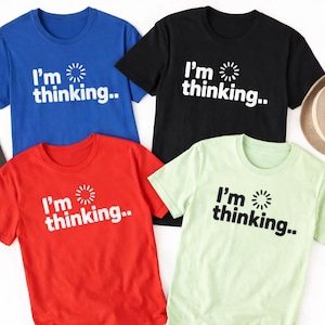 Pode incluir: Quatro camisetas de manga curta em azul, preto, vermelho e verde claro. Cada camisa tem o texto branco "I'm thinking.." com um gráfico de sol. As camisas são exibidas em uma superfície branca.