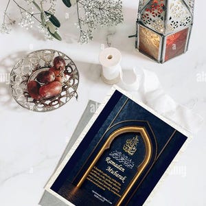 Puede incluir: Una composición de naturaleza muerta con un cuenco plateado con dátiles, una linterna decorativa con paneles rojos y dorados, y una tarjeta de felicitación con el texto "Ramadan Mubarak". La tarjeta tiene un diseño de arco dorado sobre fondo azul marino.
