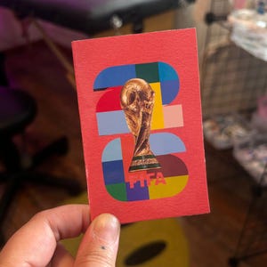 Puede incluir: Una tarjeta roja con el diseño del trofeo de la Copa Mundial de la FIFA. La tarjeta presenta un patrón geométrico colorido y el trofeo. El logotipo de la FIFA está en la parte inferior. La tarjeta se sostiene en una mano.
