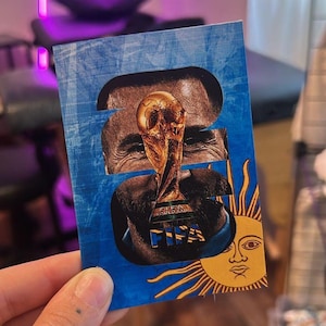 May include: A rectangular card featuring a collage of images. The card has a blue background with a golden sun and a face. The center shows the FIFA World Cup trophy and the word FIFA.