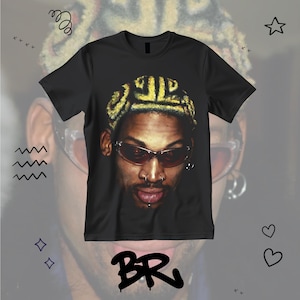 May include: Black t-shirt featuring a close-up portrait of a person wearing sunglasses and a gold-colored head covering. The image has a stylized design with decorative elements in the background.