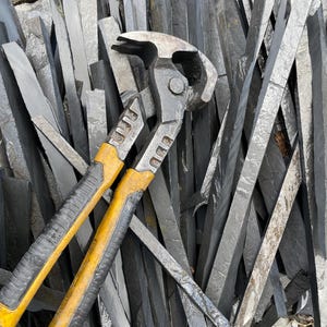 May include: A pair of yellow and black handled nippers rests atop a pile of dark gray slate pieces. The nippers have a metal head and are designed for cutting or shaping materials. The slate pieces are various sizes and shapes.