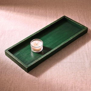 Può includere: Un vassoio rettangolare in legno verde scuro con una piccola candela accesa in un portacandele di vetro trasparente. Il vassoio ha una finitura anticata e la fiamma della candela proietta un bagliore caldo. Il vassoio è progettato per scopi decorativi.