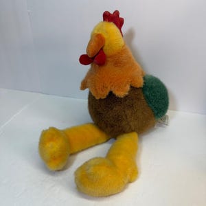 May include: A plush chicken toy featuring a yellow head, a red comb and wattle, an orange beak, a brown body, a green tail, and long yellow legs. The stuffed animal has a soft, fuzzy texture and is positioned in a seated posture.