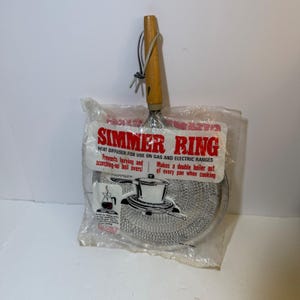 May include: A metal simmer ring heat diffuser with a wooden handle, enclosed in clear plastic packaging. The packaging features red and black text reading SIMMER RING, HEAT DIFFUSER-FOR USE ON GAS AND ELECTRIC RANGES, and includes a graphic illustration.
