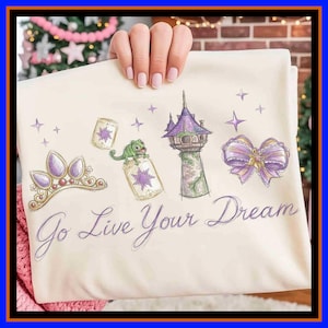 May include: A cream-coloured t-shirt featuring a purple and gold graphic print of a tiara, a green chameleon on a lantern, a tall tower, and a bow. The text "Go Live Your Dream" is printed in purple cursive script below the illustrated icons.