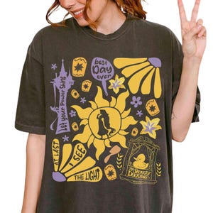 May include: Dark grey t-shirt featuring a yellow and purple graphic. The design includes a sun, flowers, and text that reads "Let your Power Shine", "At Last I See The Light", "Best Day Ever", and "The Snuggly Duckling".