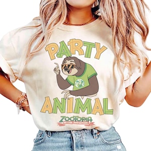 May include: A cream-coloured t-shirt featuring the words "PARTY ANIMAL" in orange and green block letters. The design includes a cartoon sloth wearing sunglasses and a green shirt. The shirt also has the text "ZOOTOPIA BETTER ZOOOGETHER".