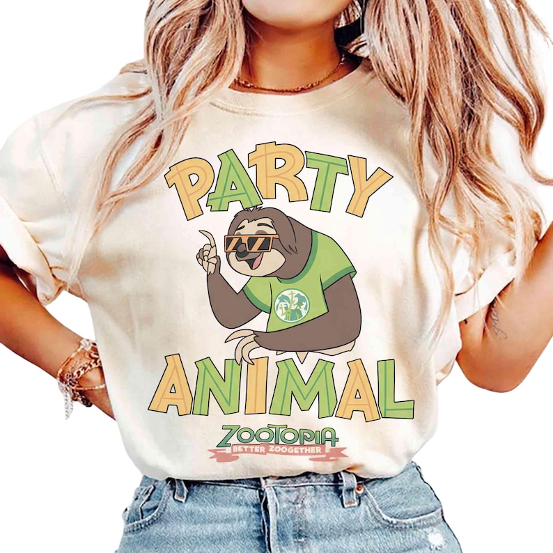 May include: A cream-coloured t-shirt featuring the words "PARTY ANIMAL" in orange and green block letters. The design includes a cartoon sloth wearing sunglasses and a green shirt. The shirt also has the text "ZOOTOPIA BETTER ZOOOGETHER".