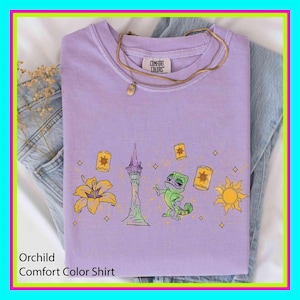 Könnte beinhalten: Orchideenfarbenes Comfort Colors-Shirt mit einer Grafik, die eine Blume, einen Turm, ein Chamäleon, Laternen und eine Sonne zeigt. Eine goldene Halskette liegt auf dem gefalteten Shirt, das auf einer Jeans liegt.