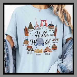 May include: Pale blue t-shirt featuring the words "Hello World" in a decorative font, surrounded by illustrations of famous landmarks. The design includes greetings in multiple languages, such as "Bonjour!" and "Ciao!"