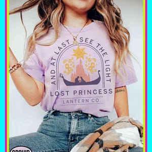 Könnte beinhalten: Ein helllila T-Shirt mit dem Text "AND AT LAST SEE THE LIGHT" und "LOST PRINCESS LANTERN CO." zeigt eine Grafik eines Bootes, einer Burg und der Sonne. Das Shirt wird mit einer blauen Jeans und einem gemusterten Schal getragen.