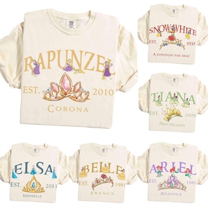 May include: Cream-colored t-shirts with various princess-themed designs. Each shirt features a princess's name, a crown, and the year of establishment. The designs include Rapunzel, Snow White, Tiana, Elsa, Belle, and Ariel.