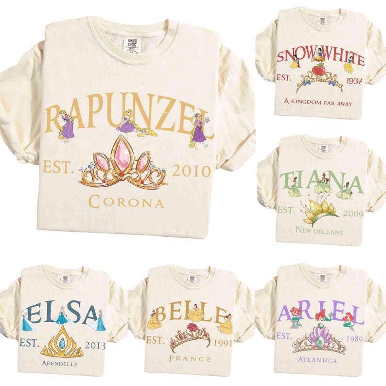 May include: Cream-colored t-shirts with various princess-themed designs. Each shirt features a princess's name, a crown, and the year of establishment. The designs include Rapunzel, Snow White, Tiana, Elsa, Belle, and Ariel.