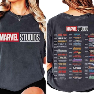 May include: Dark gray t-shirt featuring the "MARVEL STUDIOS" logo in red and white on the front. The back of the shirt lists Marvel movie titles and release years from 2008 to 2026. A comfortable, casual tee for fans of the Marvel Cinematic Universe.