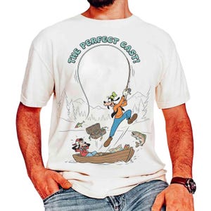 Puede incluir: Camiseta blanquecina con una ilustración de dibujos animados de Goofy pescando. Goofy está representado saltando con una caña de pescar, mientras que otro personaje está en un bote. El texto "THE PERFECT CAST!" se arquea sobre la escena.