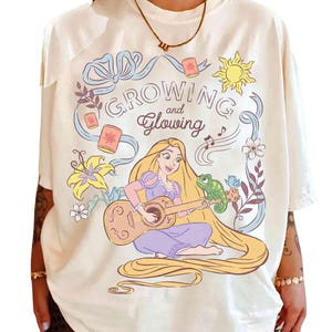 May include: Cream-colored t-shirt featuring a cartoon illustration of a woman playing a guitar, with a green chameleon. The design includes the text "Growing and Glowing" with floral and sun accents. The woman has long blonde hair.