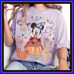 May include: A light purple t-shirt featuring a graphic of a cartoon mouse wearing a blue wizard hat with a moon and stars, sitting on a large orange pumpkin. The design includes autumn leaves, a broom, and small star accents in brown and orange.