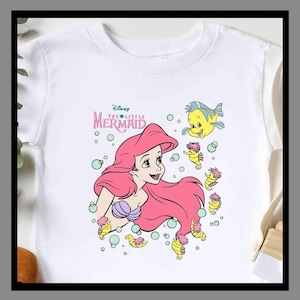 May include: White t-shirt featuring a colorful graphic of Ariel from Disney's The Little Mermaid. Ariel has long pink hair and a purple seashell top. The design includes yellow seahorses and the text "The Little Mermaid".