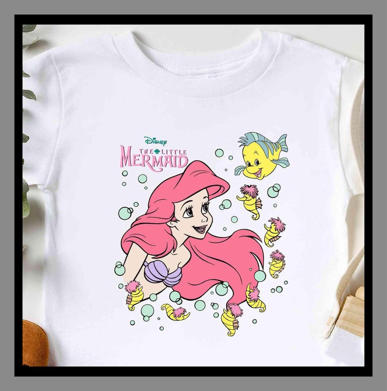 May include: White t-shirt featuring a colorful graphic of Ariel from Disney's The Little Mermaid. Ariel has long pink hair and a purple seashell top. The design includes yellow seahorses and the text "The Little Mermaid".