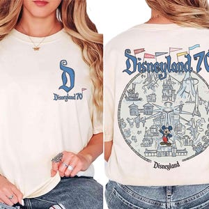 Puede incluir: Camiseta color crema con "Disneyland 70" estampada en la parte delantera y trasera. La parte delantera presenta un logotipo "D" azul y el texto. La parte trasera muestra un gr&aacute;fico circular con monumentos de Disneyland y Mickey Mouse.
