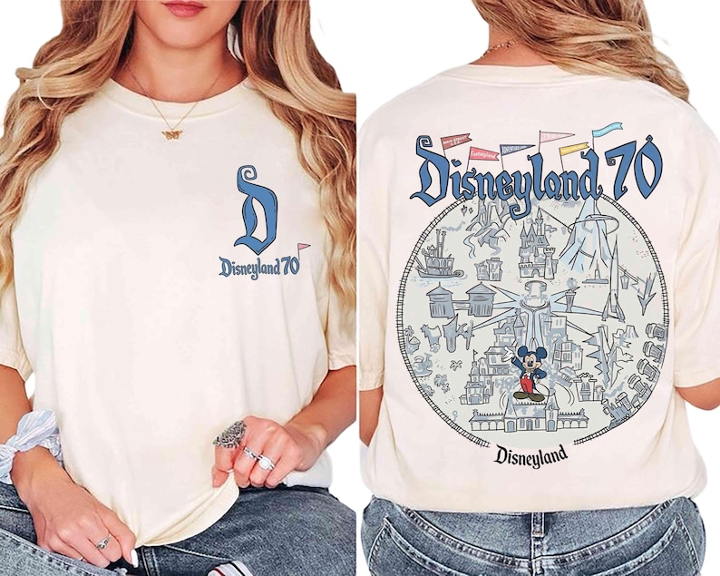 Puede incluir: Camiseta color crema con "Disneyland 70" estampada en la parte delantera y trasera. La parte delantera presenta un logotipo "D" azul y el texto. La parte trasera muestra un gr&aacute;fico circular con monumentos de Disneyland y Mickey Mouse.
