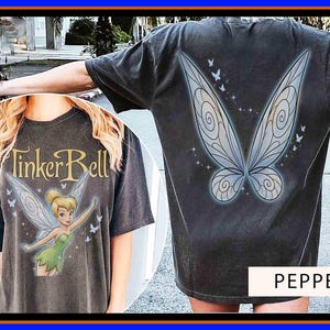 Pode incluir: Uma t-shirt cinzento-carvão com um gráfico da Sininho e o texto Tinker Bell na frente, e grandes asas de fada com pequenas borboletas e brilhos nas costas. A t-shirt está etiquetada com o nome da cor Pepper.