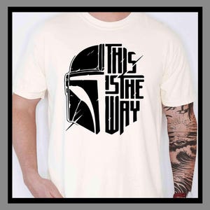 May include: Cream-colored t-shirt featuring a black graphic of a helmet and the text "THIS IS THE WAY." The design is a bold, stylized representation. The shirt is made of a soft material.