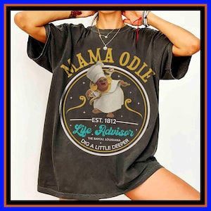Op de afbeelding: Een antracietgrijs T-shirt met korte mouwen en een cirkelvormige afbeelding met de tekst MAMA ODIE, EST. 1812, Life Advisor, THE BAYOU, LOUISIANA en DIG A LITTLE DEEPER. Het midden toont een stripfiguur in een witte mantel en hoed.