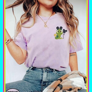 Könnte beinhalten: Ein helllila T-Shirt mit einem Cartoon-Frosch-Design, komplett mit Mickey Mouse Ohren und einer Sonne. Der Frosch ist grün mit schwarzen Details. Das Wort "ORCHID" befindet sich in einem weißen Oval unten links.