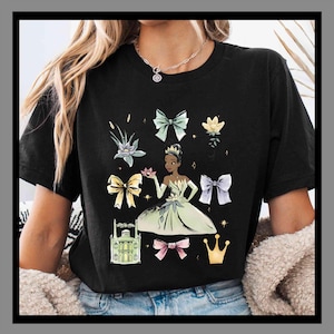 May include: Black t-shirt featuring a princess graphic with bows, flowers, a crown, and a building. The princess is wearing a green dress and holding a flower. The design includes various bows in different colors.