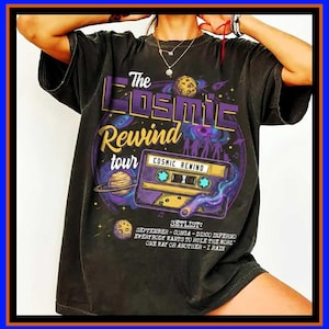 Op de afbeelding: Een zwart t-shirt met korte mouwen en een grafische print met de tekst The Cosmic Rewind tour, een cassettebandje, planeten en een setlist met September, Conga, Disco Inferno, Everybody Wants to Rule the World, One Way or Another en I Rain.