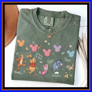 May include: An olive green crew neck t-shirt featuring a graphic print of Winnie the Pooh characters including Tigger, Piglet, Eeyore, and Roo, along with colorful Mickey-shaped balloons, honey pots, and the names of the characters in white script.
