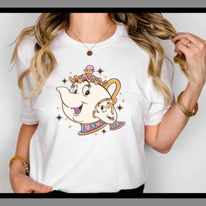 Puede incluir: Camiseta blanca con una ilustración de dibujos animados de la Sra. Potts y Chip de La Bella y la Bestia. El diseño incluye la tetera y la taza con caras sonrientes, rodeadas de estrellas. La camiseta es de cuello redondo clásico.