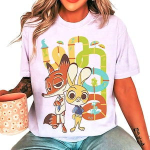 May include: A light purple t-shirt featuring a cartoon illustration of Nick Wilde and Judy Hopps from Zootopia. The design includes a colorful, whimsical background with the word "Zootopia" in a playful font.