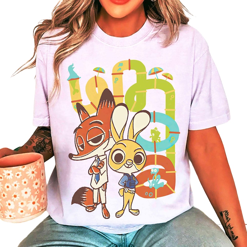May include: A light purple t-shirt featuring a cartoon illustration of Nick Wilde and Judy Hopps from Zootopia. The design includes a colorful, whimsical background with the word "Zootopia" in a playful font.