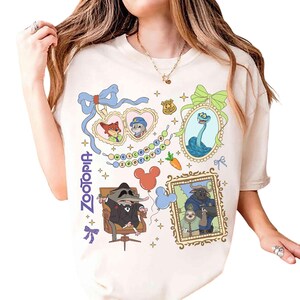 May include: Cream-coloured t-shirt with colourful Zootopia character graphics. The design includes framed images of characters, the word "Zootopia", and other decorative elements. The shirt has a crew neck and short sleeves.