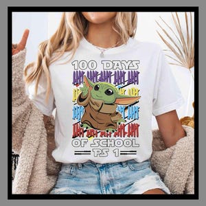 May include: White t-shirt featuring a cartoon character with the text "100 Days of School PS 1". The design includes colorful vertical lines and the character is in a brown cloak. The shirt is worn by a person.