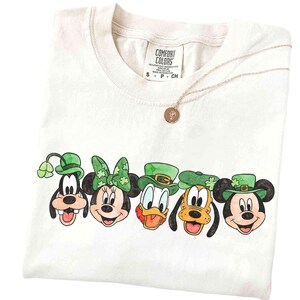 Op de afbeelding: Crèmekleurig T-shirt met gezichten van stripfiguren met groene St. Patrick's Day-hoeden. De personages zijn Goofy, Minnie Mouse, Donald Duck, Pluto en Mickey Mouse. Een gouden ketting met een hanger is ook zichtbaar.