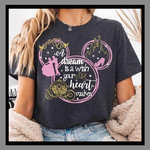 Puede incluir: Camiseta gris oscuro con un diseño rosa y dorado. El diseño presenta el texto "A dream is a wish your heart makes" e incluye una pareja bailando, un castillo, un zapato de cristal y un carruaje.