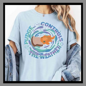 May include: Pale blue t-shirt featuring a circular design with the text "Pudge Controls The Weather." The graphic shows an orange creature and a brown and tan object within a swirling pattern with blue, purple, and green hues.