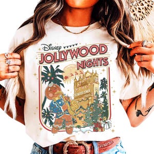 May include: Cream-colored t-shirt featuring the text "Disney Jollywood Nights" in red and gold. The graphic includes a gingerbread man playing a saxophone, palm trees, and the Hollywood Tower Hotel, with Christmas decorations.