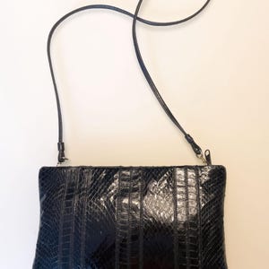 May include: Black rectangular clutch with a long, thin strap. The clutch has a textured, reptile-like pattern with vertical stitched details. The strap is attached to the top corners of the bag with small metal clasps.