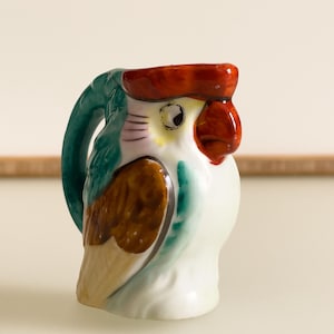 May include: A small, ceramic parrot-shaped pitcher. The pitcher features a red crest, a brown wing, and a green handle. The body is white with yellow and black details. The beak is red, and the eye is black. The pitcher is a decorative item.