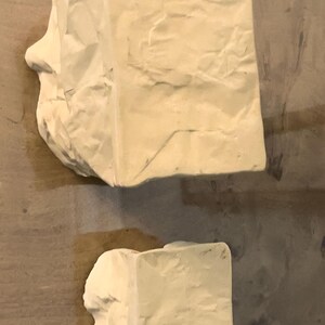 May include: Two abstract sculptures in a light beige color. The larger sculpture has a rectangular shape with a side profile of a face. The smaller sculpture is also rectangular. Both have a textured, crumpled paper appearance.