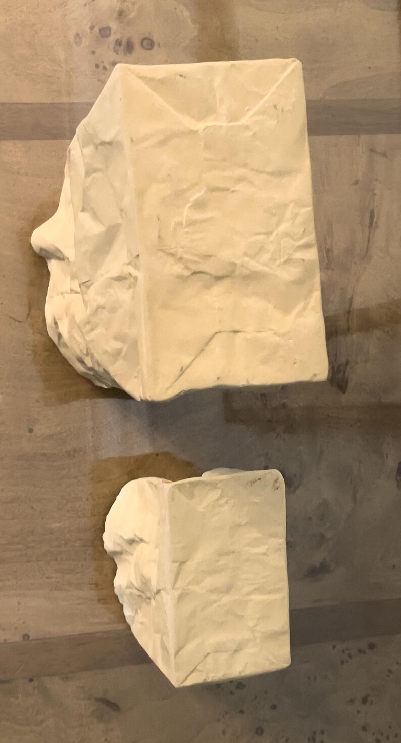 May include: Two abstract sculptures in a light beige color. The larger sculpture has a rectangular shape with a side profile of a face. The smaller sculpture is also rectangular. Both have a textured, crumpled paper appearance.