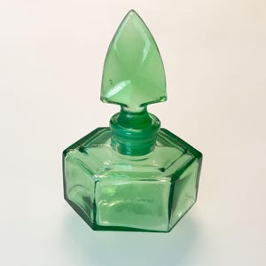 Op de afbeelding: Een doorschijnende groene glazen fles met een zeshoekige basis en een bijpassende puntige, speervormige glazen stop. Het vat heeft strakke, geometrische lijnen en een glad, gepolijst oppervlak dat licht door het levendige smaragdgroene glas reflecteert.