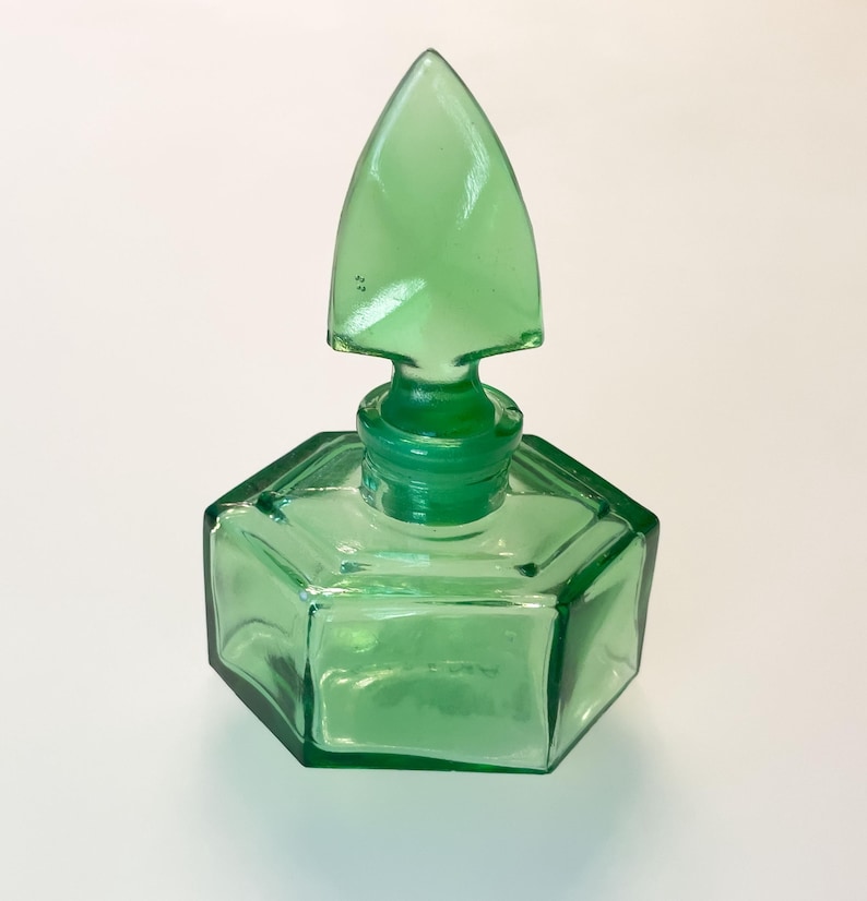 Op de afbeelding: Een doorschijnende groene glazen fles met een zeshoekige basis en een bijpassende puntige, speervormige glazen stop. Het vat heeft strakke, geometrische lijnen en een glad, gepolijst oppervlak dat licht door het levendige smaragdgroene glas reflecteert.